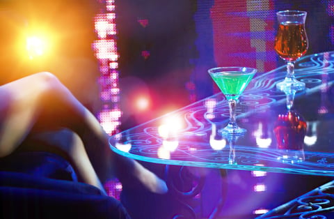 Close-up shot in dimly lit nightclub: table with green and orange cocktails under neon lights, persons legs in foreground